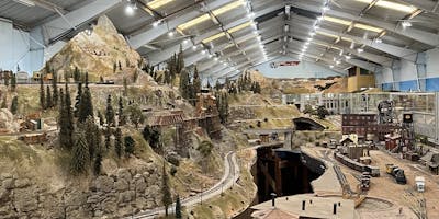 A multi-level model railway layout in a well lit warehouse, with a huge model mountain on the left hand side