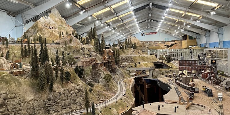 A multi-level model railway layout in a well lit warehouse, with a huge model mountain on the left hand side