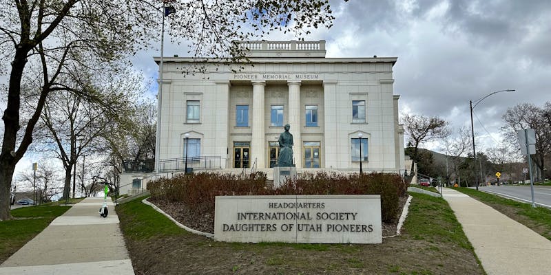 A grand building with Pioneer Memorial Museum written in gold letters along the top, and a plaque in front of it reading Headquarters International Society Daughters of Utah Pioneers