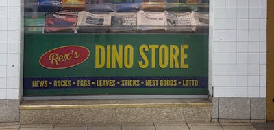 Green Rex's Dino Store sign with yellow lettering advertising NEWS • ROCKS • EGGS • LEAVES • STICKS • NEST GOODS • LOTTO, beneath a window displaying dinosaur-themed newspapers and candy bars, framed by white subway tiles.