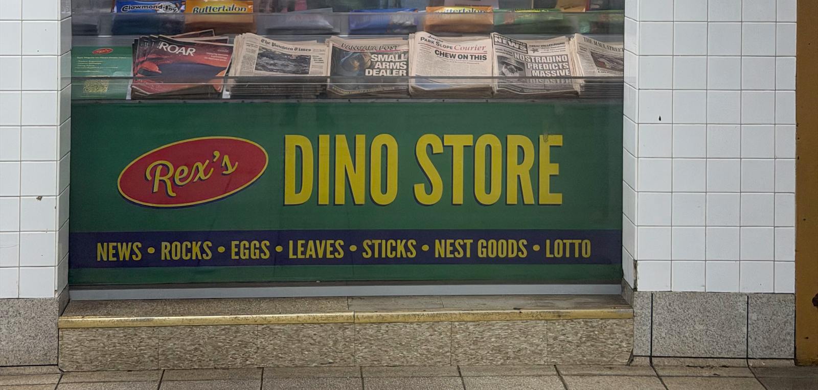 Green Rex's Dino Store sign with yellow lettering advertising NEWS • ROCKS • EGGS • LEAVES • STICKS • NEST GOODS • LOTTO, beneath a window displaying dinosaur-themed newspapers and candy bars, framed by white subway tiles.