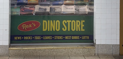 Green Rex's Dino Store sign with yellow lettering advertising NEWS • ROCKS • EGGS • LEAVES • STICKS • NEST GOODS • LOTTO, beneath a window displaying dinosaur-themed newspapers and candy bars, framed by white subway tiles.